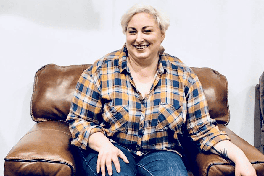Founder Andrea Kelly sits in a comfy brown leather chair while speaking on a panel. She wears a bronze and navy colored plaid shirt with the sleeves rolled up, and dark denim jeans.