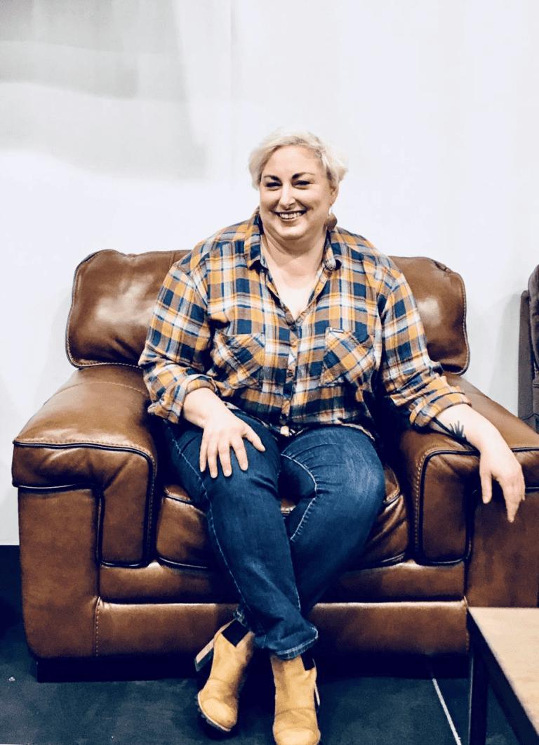 Founder Andrea Kelly, wearing a gold and blue flannel shirt and dark jeans, sits in a padded leather chair during a panel discussion. She smiles as she engages with the audience.