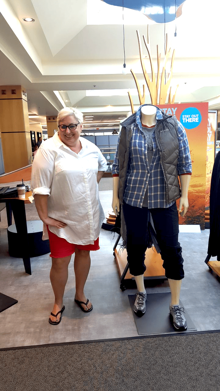 Founder Andrea Kelly stands, smiling, in a white long-sleeved button down shirt, red shorts, and sandals, next to a size 18 female mannequin dressed in a long-sleeved grey plaid shirt, a grey woven vest, black capri pants and hiking boots manufactured in Plus sizes by Columbia Sportswear.