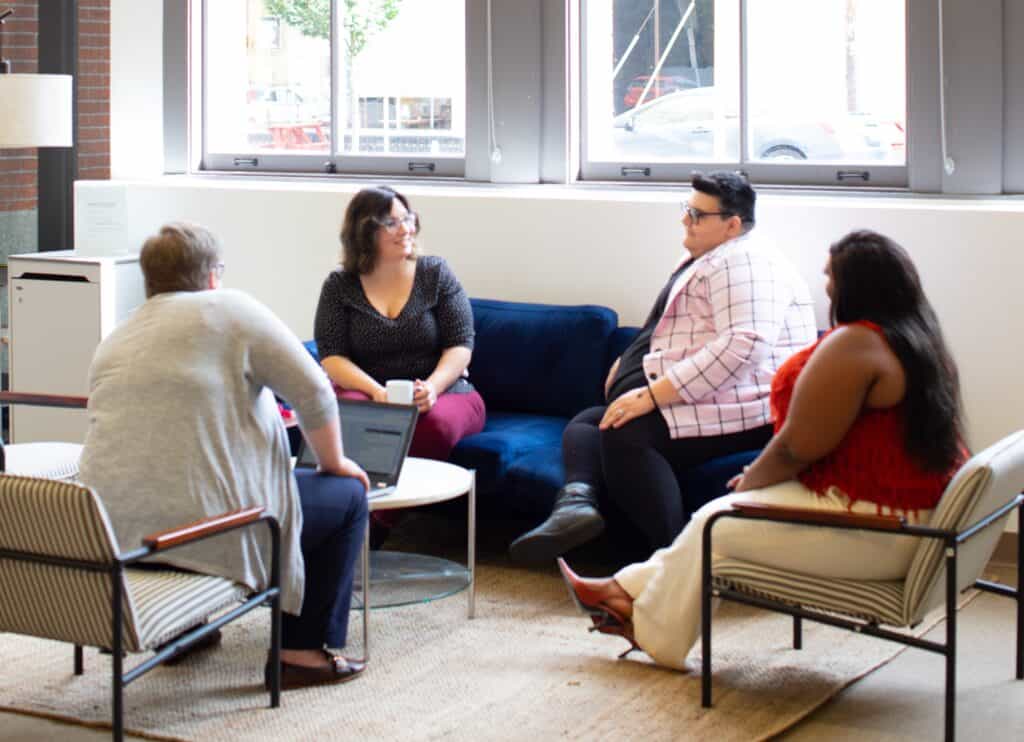 A photo of 4 people varying in size, gender identity, and ethnicity, in a well-lit business setting, chairs circled in discussion.