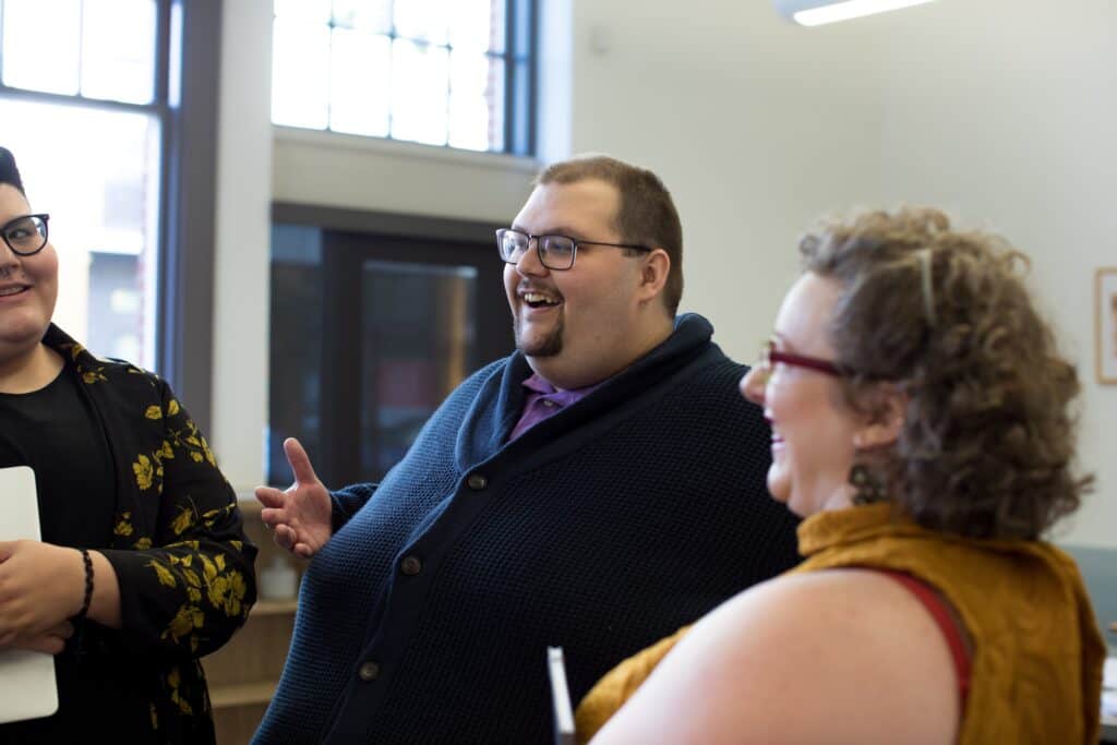A photo of 3 people varying in size, gender identity, and ethnicity, standing in a circle in a well-lit business setting, having an engaging conversation.