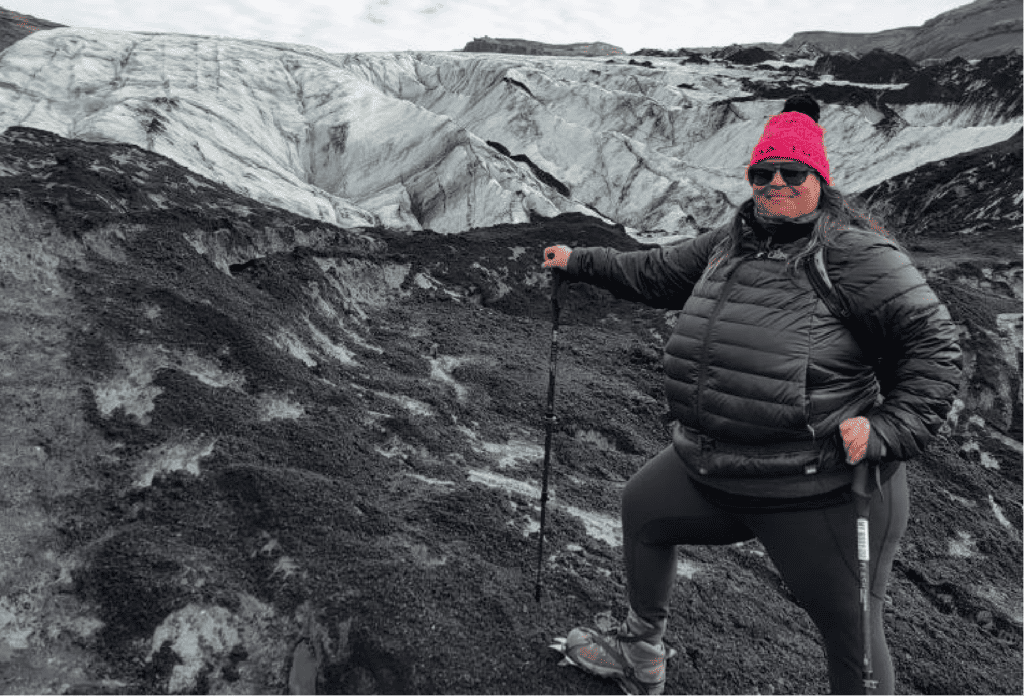 A female plus-sized hiker stands in the foreground, wearing a red beanie, sunglasses, a black puffer, black hiking pants, hiking boots and trekking poles, on a rocky alpine trail with snow-capped mountains and clouds in the background.