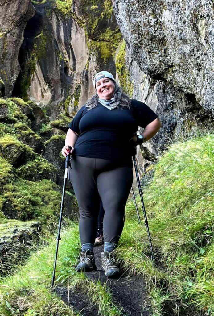 A plus-sized hiker stands, grinning, in a black short sleeved shirt, hiking pants, hiking boots. and trekking poles, on a green mossy path with craggy rocks in the background